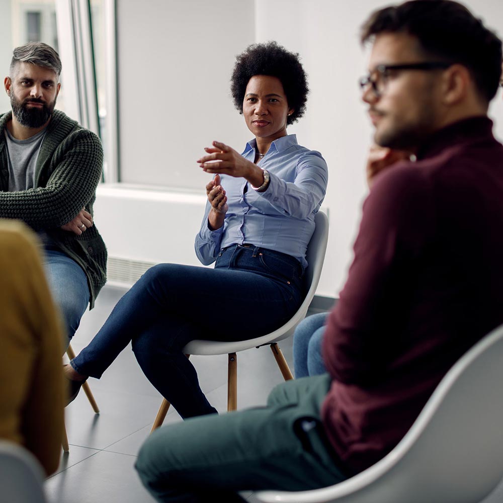 more-than-an-aftercare-plan A woman in a blue shirt gestures while speaking during a group discussion in a bright room. Three other people listen intently, creating an engaged atmosphere.
